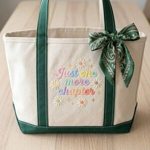 May include: Cream tote bag with green trim and handles. The bag has the embroidered text "Just one more chapter" with colourful daisy accents. A green paisley scarf is tied to the handle.