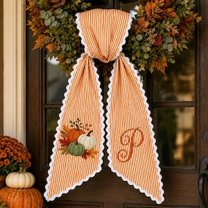May include: An orange and white striped fabric door hanger with scalloped white edges. One side features an embroidered design of three pumpkins in orange, white, and green, while the other side displays a large, embroidered cursive letter P.