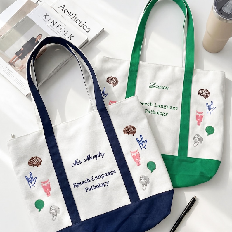 Puede incluir: Dos bolsas de lona blancas con gr&aacute;ficos bordados de patolog&iacute;a del habla. Una bolsa tiene detalles en azul marino y el texto "Mrs. Murphy Speech-Language Pathology". La otra tiene detalles en verde y el nombre "Lauren" y "Speech-Language Pathology".