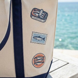 May include: A beige tote bag with navy blue trim and straps. The bag features three embroidered patches: a sardine can, a fish postage stamp, and a circular design with three fish. The background shows the ocean.