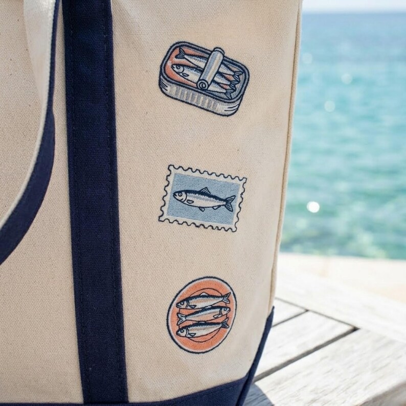 May include: A beige tote bag with navy blue trim and straps. The bag features three embroidered patches: a sardine can, a fish postage stamp, and a circular design with three fish. The background shows the ocean.