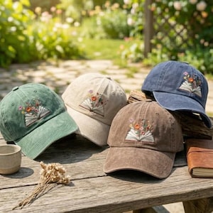 May include: Three baseball caps in sage green, beige, and brown, each featuring an embroidered design of an open book with colorful flowers emerging from the pages. The caps have a structured crown, a curved brim, and a distressed fabric finish.