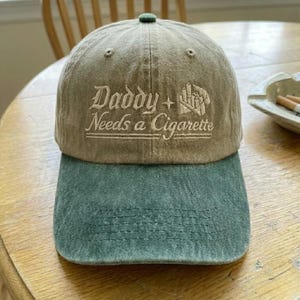 May include: A two-tone baseball cap with a khaki crown and a green bill. The cap features the embroidered text "Daddy + Needs a Cigarette" in a stylized font. The cap has a green button on top.