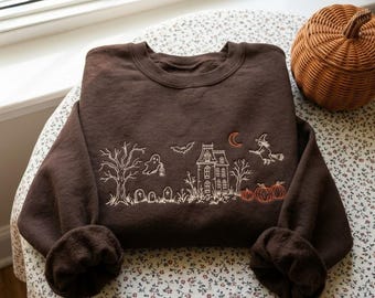 May include: A dark brown crewneck sweatshirt featuring white embroidered line art of a haunted house, a ghost, a bat, a witch on a broomstick, and bare trees. The design includes a small orange crescent moon and three orange pumpkins.
