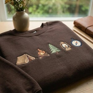 Peut inclure: Un sweat-shirt &agrave; col rond marron fonc&eacute; orn&eacute; de broderies repr&eacute;sentant une tente beige, un feu de camp aux flammes orange, un sapin vert, une lanterne beige et une boussole bleue et blanche align&eacute;s horizontalement sur la poitrine.