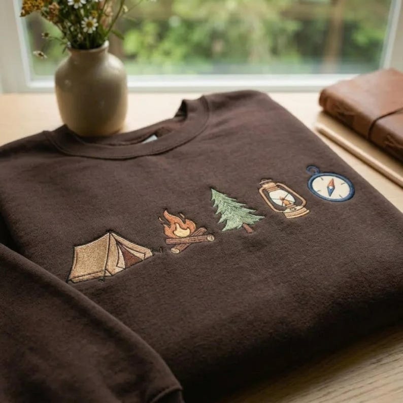 Peut inclure: Un sweat-shirt &agrave; col rond marron fonc&eacute; orn&eacute; de broderies repr&eacute;sentant une tente beige, un feu de camp aux flammes orange, un sapin vert, une lanterne beige et une boussole bleue et blanche align&eacute;s horizontalement sur la poitrine.