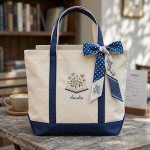 May include: A cream-colored canvas tote bag with navy blue trim and handles. The front features an embroidered design of an open book with flowers emerging from the pages, with the name Amelia embroidered below. A blue polka-dot ribbon is tied.