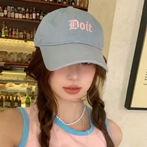 May include: A light blue baseball cap with the word "Doit" embroidered in pink script. The cap has a curved brim and a classic six-panel design. The hat is made of a soft, durable fabric.