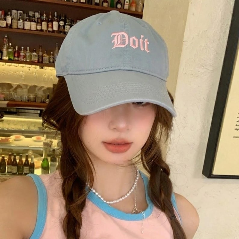 May include: A light blue baseball cap with the word "Doit" embroidered in pink script. The cap has a curved brim and a classic six-panel design. The hat is made of a soft, durable fabric.