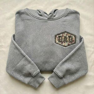 May include: A gray hooded sweatshirt with the word "DAD" embroidered on a camouflage patch. The sweatshirt has long sleeves and a drawstring hood. The fabric appears to be a soft, comfortable material.