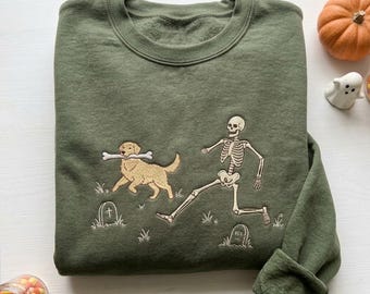 Puede incluir: Una sudadera de cuello redondo color verde oliva con un diseño bordado de un perro golden retriever llevando un hueso en la boca mientras corre junto a un esqueleto humano en un cementerio con lápidas pequeñas y matas de hierba.