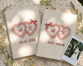 Puede incluir: Dos libros de votos de boda de lino blanquecino con corazones e iniciales bordados en rojo. Un libro dice "09.15.2026" y el otro dice "our vows". Una pequeña foto de una pareja está al lado de los libros.
