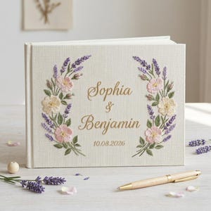May include: A cream-colored wedding guest book with embroidered floral wreath design. The wreath features pink and cream flowers with purple lavender. The names "Sophia & Benjamin" and the date "10.08.2026" are embroidered in gold.