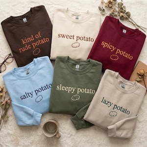 May include: Six crewneck sweatshirts in various colors, each featuring embroidered text and a small potato graphic. The shirts are dark brown, cream, maroon, light blue, olive green, and beige, with phrases like kind of rude potato, sweet potato, spicy potato, salty potato, sleepy potato, and lazy potato.