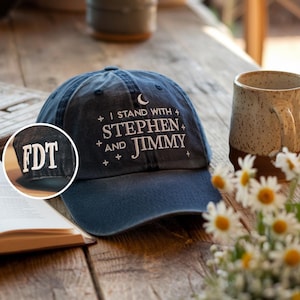 May include: A navy blue baseball cap with white embroidered text that reads "I STAND WITH STEPHEN AND JIMMY." The cap also features a crescent moon and stars design. A smaller inset shows "FDT" embroidered on the side.