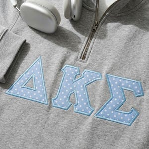 May include: A heather gray quarter-zip sweatshirt featuring the Greek letters Delta, Kappa, and Sigma in light blue fabric with a white polka dot pattern. The letters are appliqued with a light blue stitched border. Silver headphones are visible.