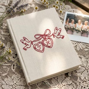 May include: A cream-colored wedding guest book with red embroidered doves, hearts, and initials. The book is rectangular and lies open on a lace tablecloth, with a framed photo of bridesmaids in the background. The cover features a decorative design.