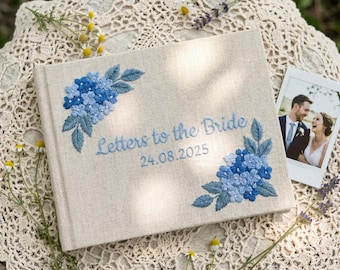 Könnte beinhalten: Ein beiges Leinenbuch mit blauen, gestickten Blumenmotiven und dem Text "Letters to the Bride 24.08.2025". Neben dem Buch befindet sich ein kleines Foto eines Paares, im Hintergrund eine Spitzendecke und Blumen.
