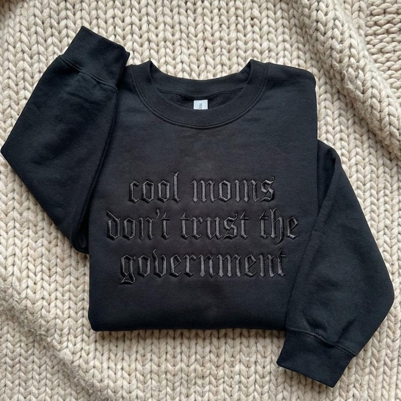 May include: Black crewneck sweatshirt with the embroidered text "cool moms don't trust the government." The sweatshirt is made of a soft material and is displayed on a neutral-colored, textured surface. The text is in a gothic font.