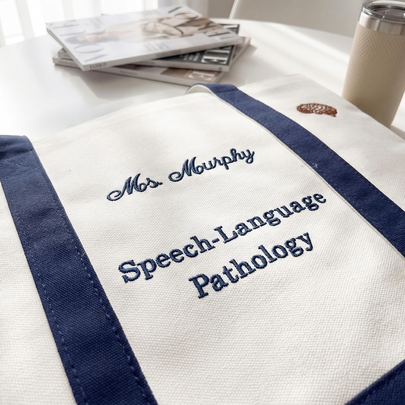 Puede incluir: Bolso tote de lona blanca con ribetes y asas azul marino. El bolso est&aacute; bordado con el nombre "Mrs. Murphy" y las palabras "Speech-Language Pathology" en hilo azul marino. Un peque&ntilde;o dise&ntilde;o bordado marr&oacute;n est&aacute; presente.