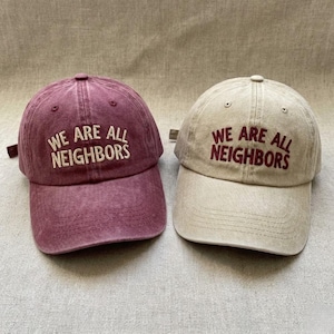 May include: Two baseball caps, one burgundy and one tan, are displayed. Both caps have the phrase "WE ARE ALL NEIGHBORS" embroidered on the front in a contrasting color. The caps have a classic six-panel design with a curved brim.