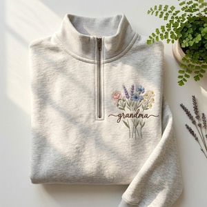 May include: A light gray heathered quarter-zip sweatshirt featuring an embroidered bouquet of colorful wildflowers and the word grandma in cursive script on the left chest. The garment has a ribbed collar and a metal zipper closure at the neck.