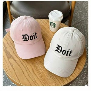 May include: Two baseball caps, one light pink and one white, sit on a wooden table. Each cap has the word "Doit" embroidered in black gothic lettering. A white paper coffee cup with a green logo is also on the table.