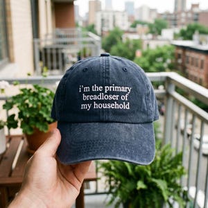 May include: A navy blue baseball cap with the text "i'm the primary breadloser of my household" in white. The cap is made of a soft material and has a curved brim. The background shows a city scene.