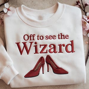 May include: A white sweatshirt with the embroidered phrase "Off to see the Wizard" in red. Below the text are two red high-heeled shoes. The sweatshirt is a crew neck style.