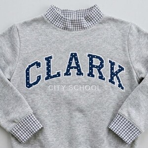 May include: A heather gray long-sleeve sweatshirt featuring a mock neck collar and sleeve cuffs in a gray and white gingham pattern. The chest displays the dark blue text CLARK in a polka dot print, with the words CITY SCHOOL embroidered below.