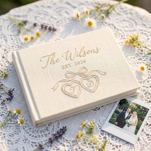 May include: A rectangular, cream-colored fabric photo album featuring gold foil lettering that reads The Wilsons EST. 2026, accompanied by a gold foil illustration of two connected hearts with the letters T and D inside and a ribbon bow above.