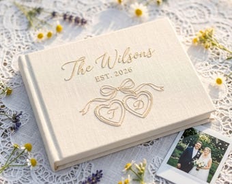 May include: A rectangular, cream-colored fabric photo album featuring gold foil lettering that reads The Wilsons EST. 2026, accompanied by a gold foil illustration of two connected hearts with the letters T and D inside and a ribbon bow above.