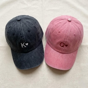 May include: Two baseball caps, one charcoal gray and one pink. The gray cap has a white "K" with a heart, and the pink cap has a "Q" with a heart. Both caps have a curved brim and a button on top.