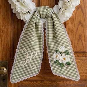 May include: A decorative wreath with white flowers and a green and white gingham bow. The bow is trimmed with white lace and features an embroidered floral design and the letter "H".