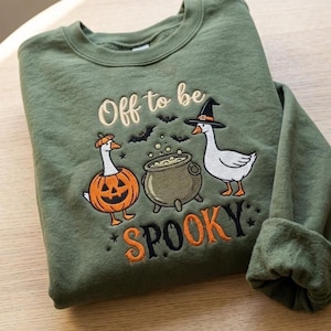 May include: An olive green crewneck sweatshirt featuring an embroidered design of two white geese, one wearing a pumpkin costume and the other a witch hat, standing beside a cauldron with the text Off to be SPOOKY and small bat silhouettes.
