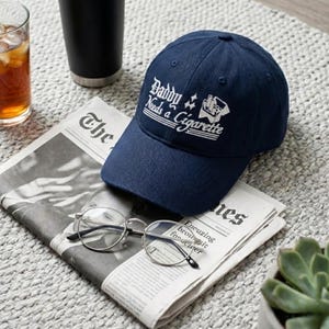 May include: A navy blue baseball cap with the text "Daddy Needs a Cigarette" in white script. The cap is resting on a newspaper with a pair of round eyeglasses. A glass of iced tea and a black tumbler are also in the image.