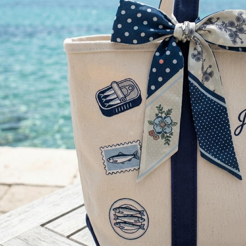 May include: Cream-colored tote bag with navy blue accents and decorative patches featuring sardines. A navy blue and white polka dot bow and floral scarf adorn the handle. The bag is on a wooden surface with a blurred ocean background.