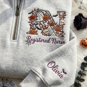 May include: A light gray quarter-zip sweatshirt featuring an embroidered letter N decorated with pumpkins, skulls, spiderwebs, and bats. Below the letter, the text Registered Nurse is embroidered in purple. The sleeve features the name Olivia.
