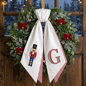 May include: A circular wreath made of green eucalyptus and cedar foliage, decorated with small red velvet bows and gold bells. A white fabric ribbon hangs from the center, featuring an embroidered nutcracker figure and the letter G.
