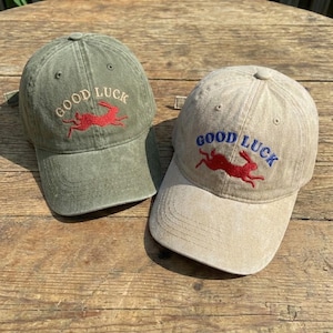 May include: Two baseball caps, one olive green and one tan, are displayed. Both caps feature the embroidered text "GOOD LUCK" above a red rabbit graphic. The olive green cap has red embroidery, while the tan cap has blue embroidery.