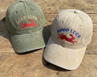 May include: Two baseball caps, one olive green and one tan, are displayed. Both caps feature the embroidered text "GOOD LUCK" above a red rabbit graphic. The olive green cap has red embroidery, while the tan cap has blue embroidery.