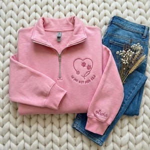 May include: A light pink quarter-zip sweatshirt with a heart-shaped design featuring paw prints and a stethoscope, with the text "In my vet med era." The sleeve is embroidered with the name "Olivia." Paired with blue jeans.