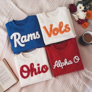 May include: Four crewneck sweatshirts in blue, white, and red. Each sweatshirt features a different word in a textured, raised font: "Rams", "Vols", "Ohio", and "Alpha O". The sweatshirts are arranged on a soft, textured surface with a book, flowers, and a mug.