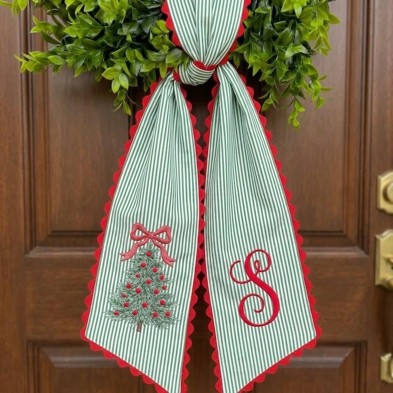 May include: A decorative fabric bow featuring a green and white vertical pinstripe pattern with red scalloped edges. One tail displays an embroidered Christmas tree with a red bow topper, while the other tail features a large red embroidered letter S.