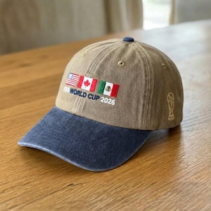 May include: A brown baseball cap with a navy blue brim. The cap features embroidered flags of the USA, Canada, and Mexico, along with the text "FIFA WORLD CUP 2026". A gold-colored FIFA World Cup trophy is embroidered on the side.