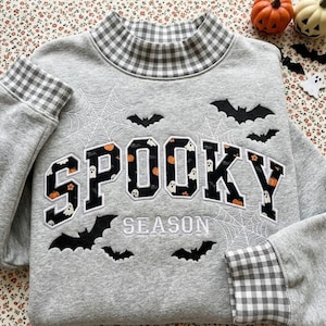 May include: A heather gray sweatshirt featuring a mock neck and cuffs with a gray and white gingham pattern. The front displays the word SPOOKY in black block letters filled with ghosts and pumpkins, with the word SEASON embroidered below.