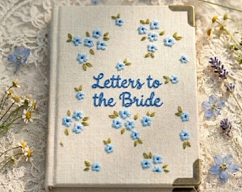 May include: A linen-covered book with the embroidered text "Letters to the Bride" in blue script. The cover is adorned with small blue floral embroidery and green leaves. The book has brass corner protectors.