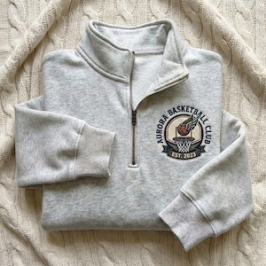 May include: A light gray sweatshirt with a quarter-zip neckline and ribbed cuffs. The sweatshirt features a circular patch with the text "AURORA BASKETBALL CLUB" around a basketball with wings and a net. The patch also includes "EST. 2023".