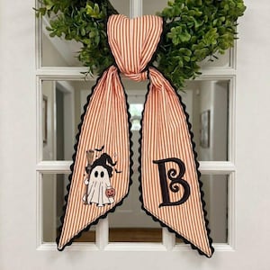 May include: A decorative fabric bow with orange and white vertical stripes and black scalloped edges. One tail features an embroidered ghost wearing a witch hat and holding a broom and pumpkin, while the other tail displays a large black letter B.