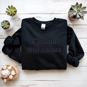 May include: A black sweatshirt with the words "Custom Sweatshirt" embroidered in a gothic font. The sweatshirt is displayed on a white wooden surface, with small potted succulents and seashells in the background.